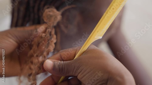 Closeup stylist combing braid ends with applicator and tube, careful squeeze and comb motion shapes extension tips, focused hands show dexterity and finishing technique under soft studio light, salon