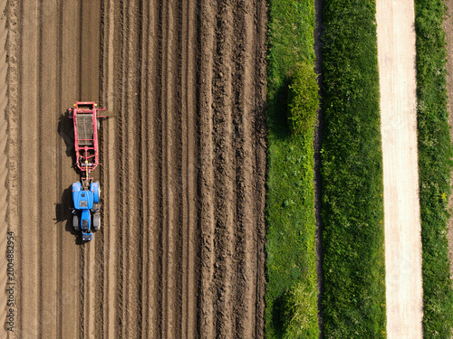 Aerial view of a blue tractor and red farm machinery working in a furrowed brown field next to a green verge and dirt road in Knottingley, England, United Kingdom.