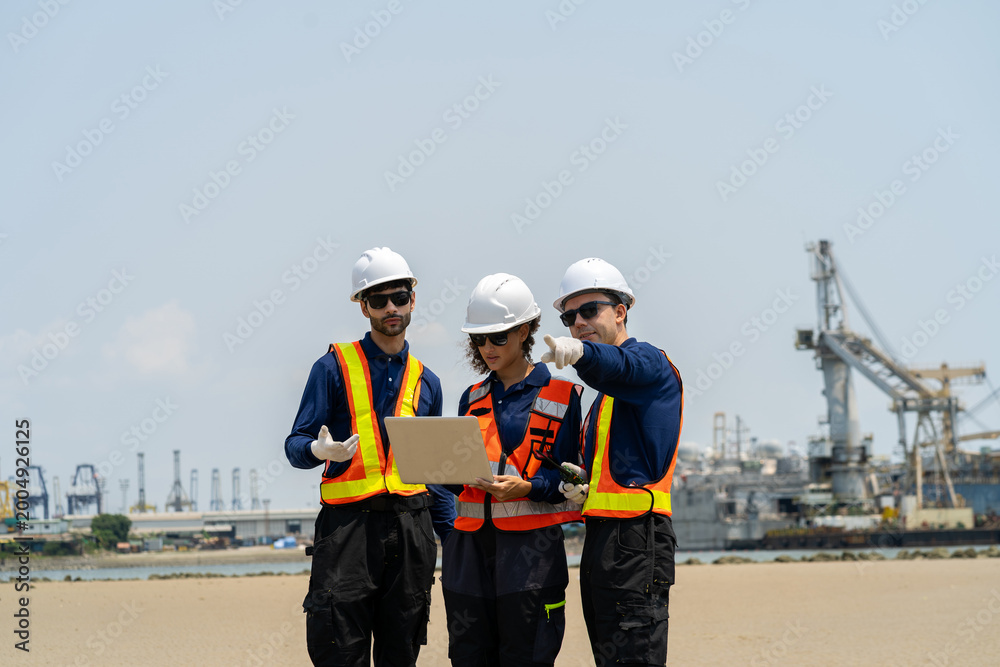Fototapeta premium A group of engineers, wearing safety suits and helmets, are examining data on laptops in a shipyard and port area, with cranes and large ships in the background.