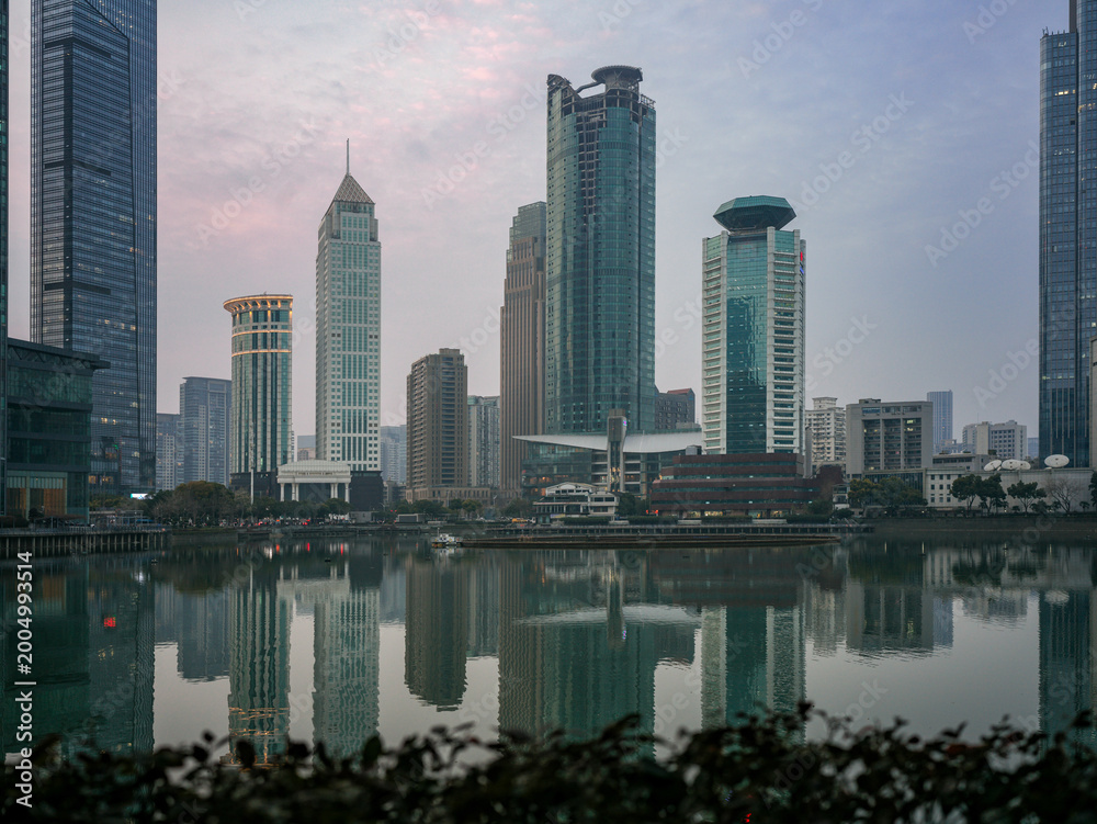 Fototapeta premium Wide twilight cityscape with modern skyscrapers reflecting in calm lake with foreground plants