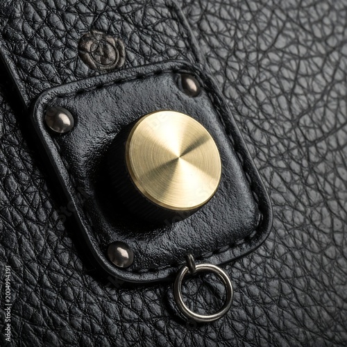 Close-up of a textured black leather surface. Features a decorative, golden-toned button and a small metal ring. Details are meticulously presented