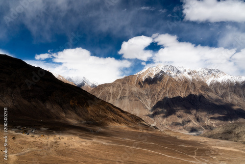 Dramatic light and shadow over snow-capped Himalayan peaks in Ladakh, India. Breathtaking mountain landscape with majestic clouds under a bright sun. Epic winter scenery of high altitude rocky desert.