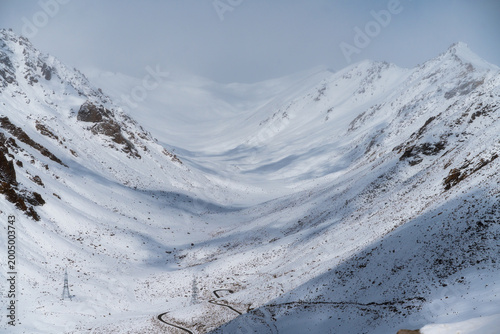 Magnificent snowy valley in Ladakh, India, with a winding road and power lines through high Himalayan peaks. Winter landscape of remote mountain pass under misty sky. Adventure and travel background.