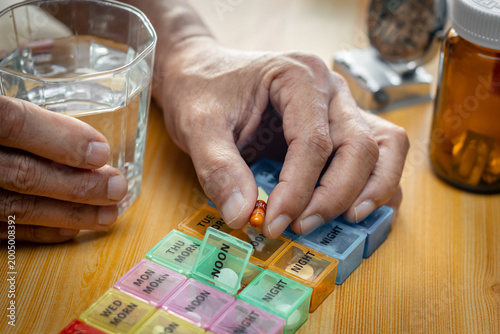 
Senior man's hands taking daily medication from a colorful pill organizer at home. Health care, medicine, and elderly wellness concept featuring a glass of water and pill bottles on a wooden table.