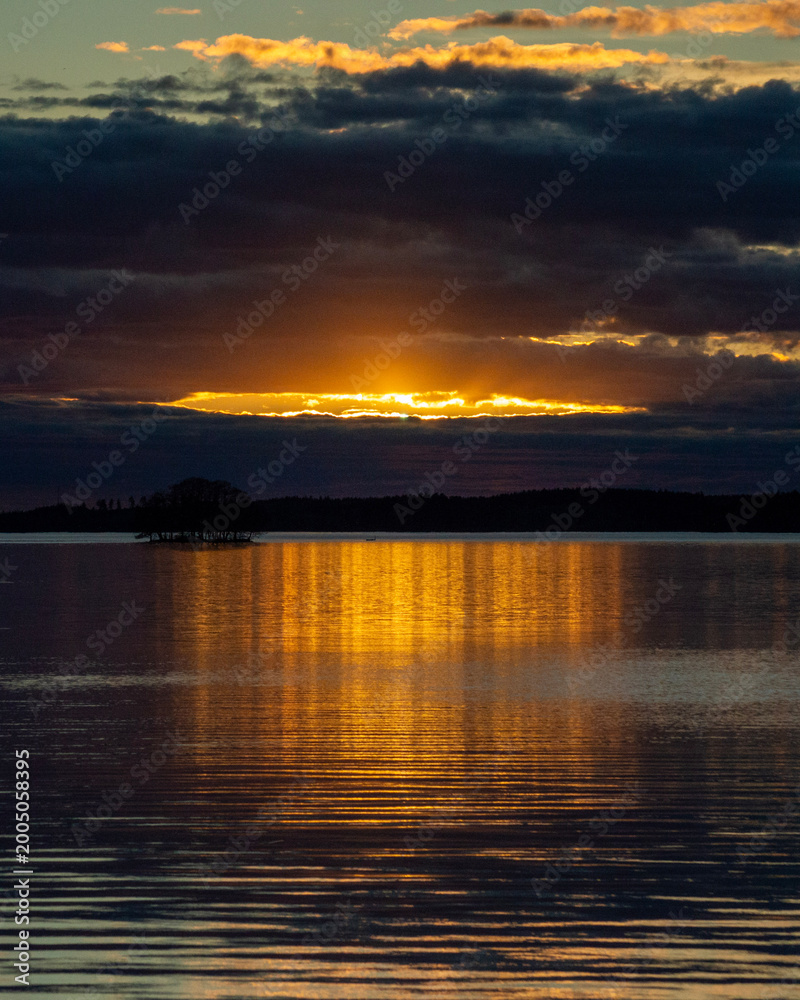 Fototapeta premium Dramatic Golden Sunset Breaking Through Dark Storm Clouds Over Finnish Lake with Island Silhouette
