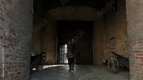  Medieval warrior running through dark arched castle gate into fortress courtyard and drawing sword. Dynamic cinematic action entry shot with cannon props — historical reenactment adventure footage