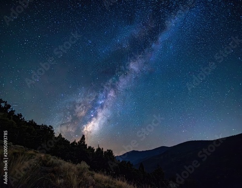 A breathtaking view of the Milky Way galaxy arches over a silhouetted mountain range and forested landscape.