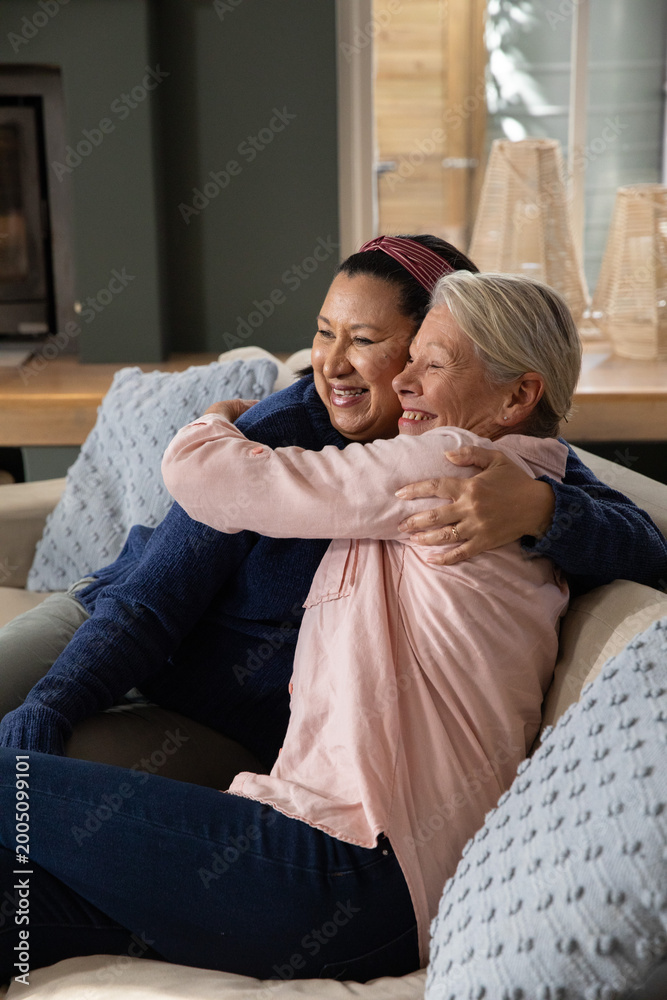 Fototapeta premium Diverse female friends wearing sweater and blouse sitting on living room sofa, hugging with pillows