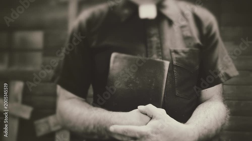 Clergyman, reverend or priest wearing a clerical collar and clutching a bible. Preacher preaching the gospel in front of an old rustic rural church or cemetery.