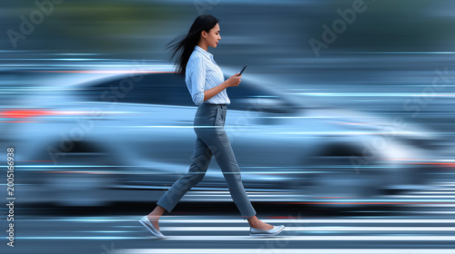 Woman walks on street while using smartphone with blurred car passing by in city environment during daytime