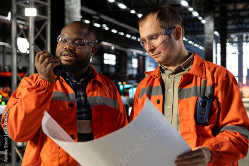 Two diverse engineers use plans to oversee construction and production tasks, surrounded by industrial machinery, metal tools and engineering elements that support plant operation.