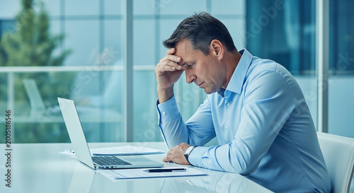 stressed businessman rubbing temples while working on laptop in bright office