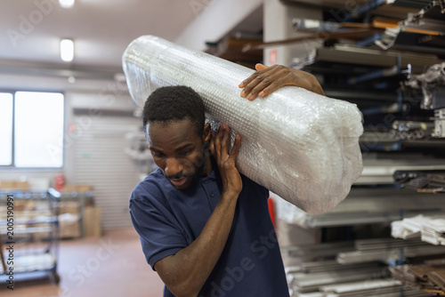 Принти на полотні Warehouse worker manually handling heavy packaged goods on his shoulder in logistics settings, having a physically demanding job