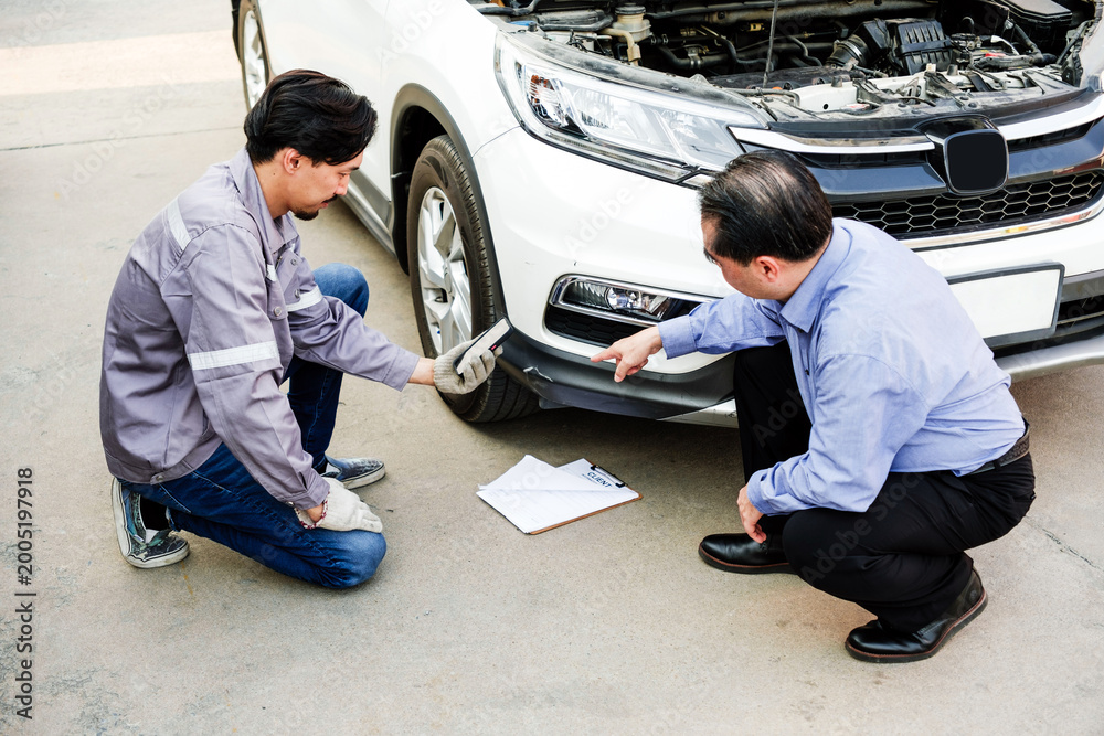 Fototapeta premium Mechanic and client kneel by a white car with the hood open, inspecting front bumper damage and discussing repairs beside a clipboard.