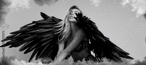 Sensual woman in black lingerie wearing decorative angel wings, crouched in a studio, black and white artistic photography.
