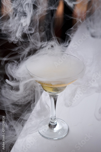 Smoking Cocktail in Coupe Glass with Dry Ice Effect. A close-up of a golden cocktail in a glass surrounded by thick white smoke or vapor from dry ice. Professional molecular mixology at a bar
