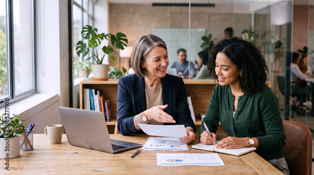 Obraz premium Smiling Senior Businesswoman Mentoring Young Mixed Race Female Colleague Over Reports and Laptop in Bright Office
