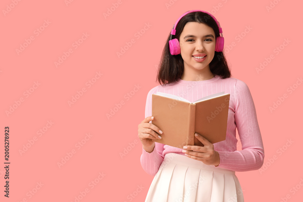 Fototapeta premium Happy young woman with book and headphones listening to audiobook on pink background