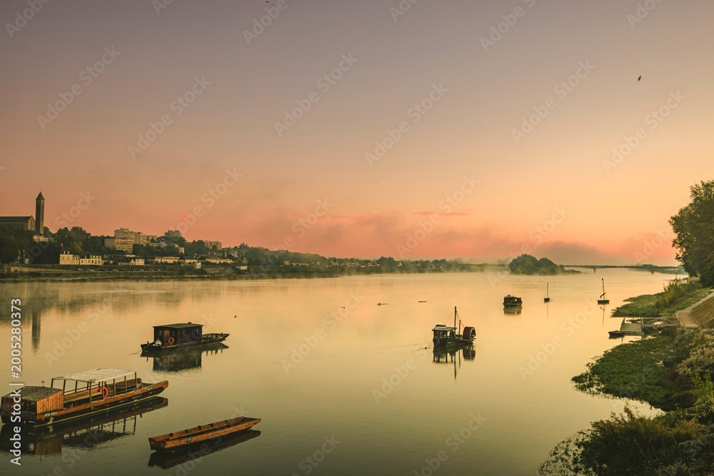 Fototapeta premium Golden hues reflect off the calm waters of the Loire River as boats gently bob in the soft evening light. Blois lies peacefully in the backdrop, blending history with stunning landscapes