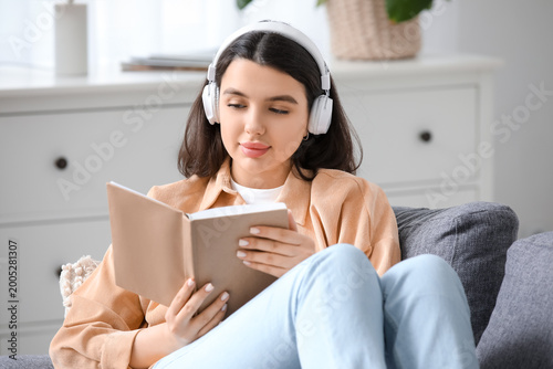 Pretty young woman with headphones and book listening to audiobook on sofa in...