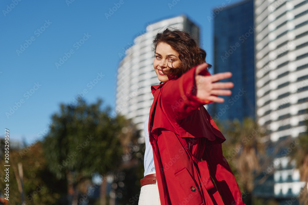 Obraz na płótnie Young woman in a red coat extends her arm toward the camera with a playful smile, standing in a bright urban setting with modern buildings, trees, and blue sky behind