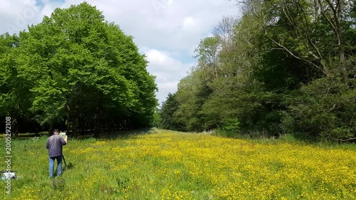 Painter standing in meadows with his easel, painting a spring landscape of trees, grass and buttercups on a sunny day.