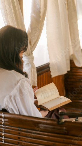 Woman in vintage clothing reading an old book by a window with curtains