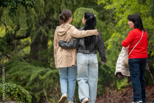 Two young women walking arm in arm through a lush green park, smiling at each other during a relaxed nature walk with soft forest background.
