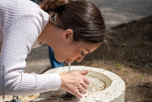 Young woman drinking fresh water from a public outdoor fountain in bright sunlight, bending over during a walk in a park or urban green space.
