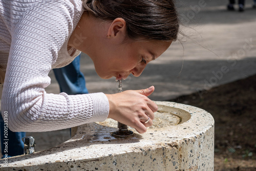 Young woman drinking fresh water from a public outdoor fountain on a sunny day, refreshing herself during a walk in a park.