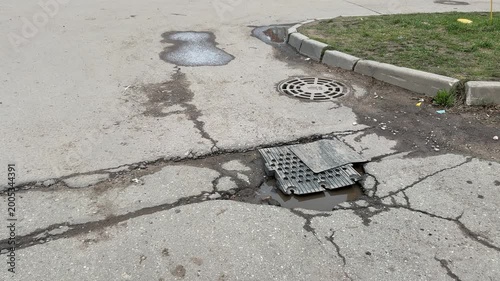 Large pothole on asphalt road covered with improvised metal plate, cracked pavement radiating fractures, shallow water pooling.