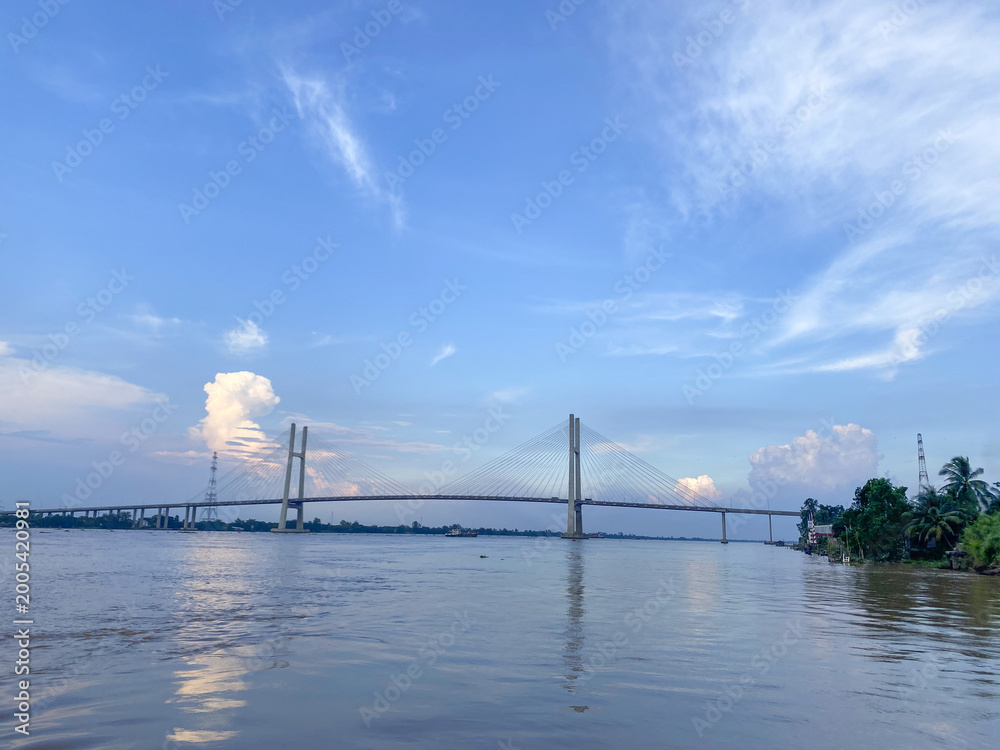 Fototapeta premium Magnificent view of a bridge spanning a wide river at sunset, with lush green landscapes and a vibrant sky, capturing urban connectivity and natural beauty.