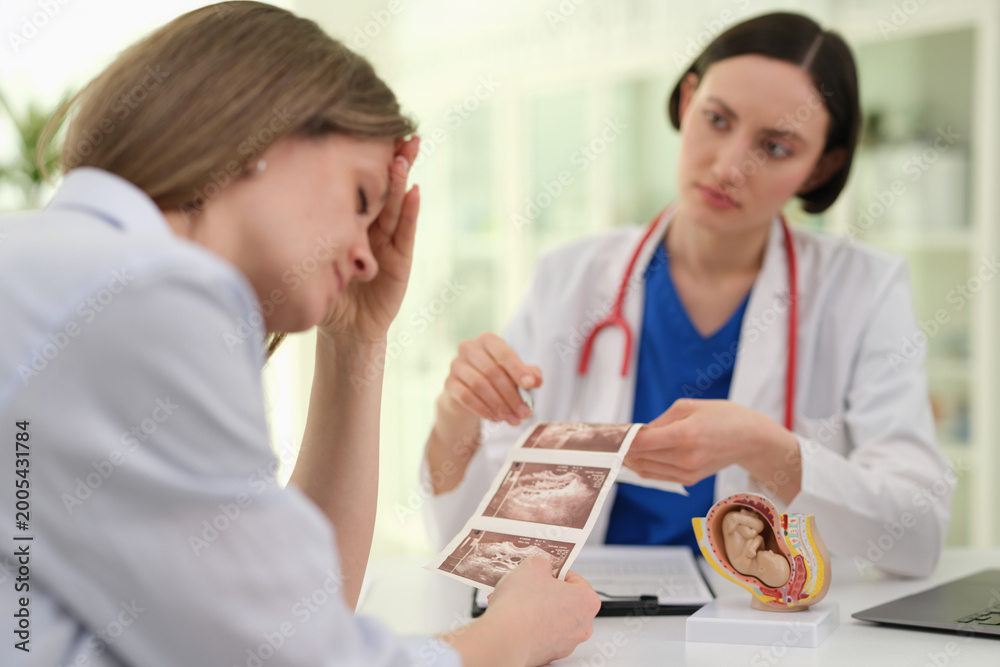 Fototapeta premium Doctor reviewing ultrasound photos with concerned patient during prenatal counseling, fetal model and charts on desk, empathetic