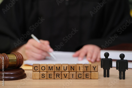 In a courtroom setting, a judge in black robes attends to paperwork related to community service. Wooden blocks spell out community service next to a gavel and figurines.