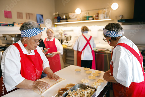 A group of diverse elderly women wearing red aprons and blue headbands work together to prepare handmade pastries in a bright kitchen