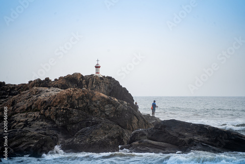 Fishermen fishing from rugged rocks along Goa coastline with waves crashing and a small coastal marker, capturing serene seaside livelihood in India.