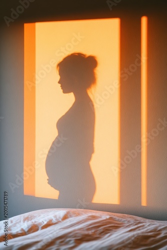 Silhouette of a pregnant woman cast as a shadow on a wall, illuminated by warm orange light, with a soft bed visible in the foreground