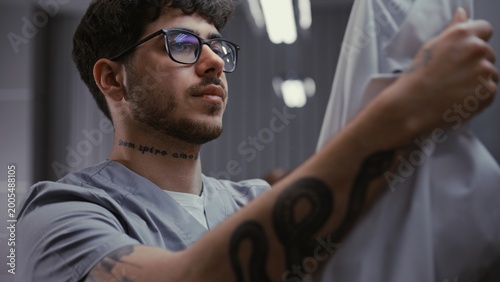 Billede på lærred Close Up of Young Male Doctor in Glasses, Removes Protective Mask, Rubs Eyes From Fatigue, and Takes Off White Medical Coat in Hospital Locker Room