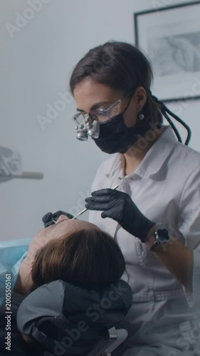 Professional female dentist wearing protective mask and magnifying glasses carefully examining patient’s teeth during modern dental checkup in bright clinic