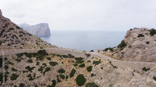 Aerial drone shot of a yellow car and cyclist on the serpentine road to Cap de Formentor lighthouse, Mallorca. Scenic curved asphalt mountain road and rocky coast in Spain. 4K