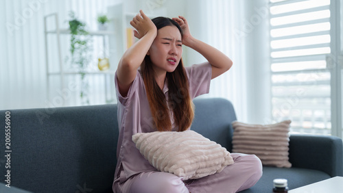 young Asian woman suffering headache migraine pain sitting on sofa holding head stressed frustrated feeling unwell depressed at home living room mental health