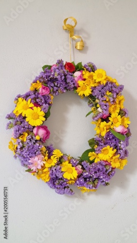 A vibrant handmade floral wreath hangs on a light-colored wall, decorated with a mix of yellow daisy-like flowers, purple statice, and pink roses