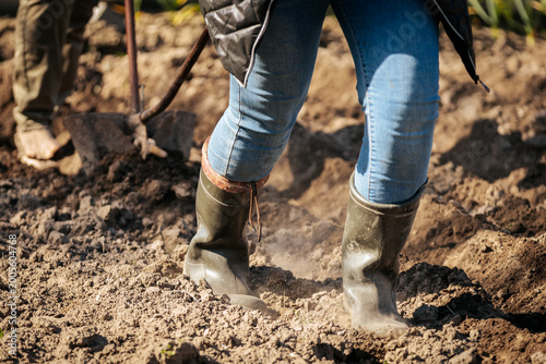 Person wearing green rubber boots and blue jeans working in a garden, soil being turned with a shovel in the background under bright sunlight