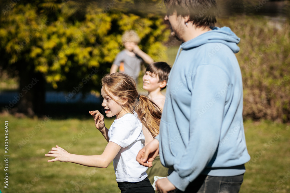 Fototapeta premium Group of children and adult running together in a park on a sunny day, with green grass and trees in the background creating a lively outdoor scene