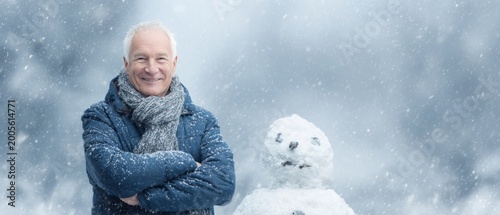 Smiling Senior Man Standing Beside Snowman During Gentle Winter Snowfall