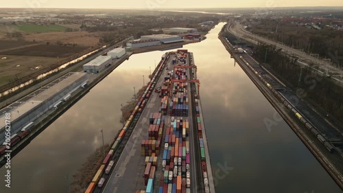 Aerial orbital drone flight circling the Gliwice inland port container terminal during sunset. View of colorful containers, cranes, and Gliwice Canal with reflections in the water.