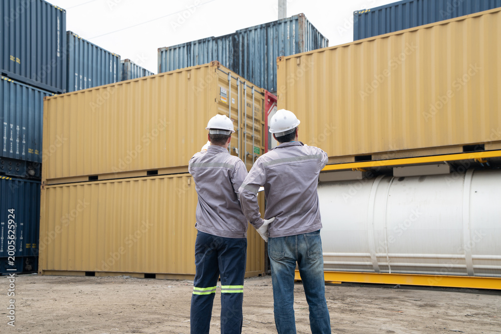 Fototapeta premium Two engineers workers in hard hats inspect stacked shipping containers at a freight yard, discussing logistics and safety.