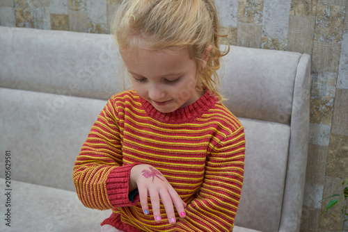 Cute little blonde girl looking at her glitter tattoo on hand ,a blonde girl in a striped sweater showing a colorful manicure and a temporary tattoo in the shape of a butterfly,