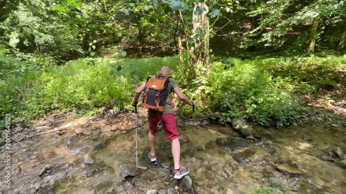 4K POV video of a man with a backpack and trekking poles crossing a shallow stream in a lush, green forest on a sunny day. Nature trekking, hiking adventure, and active lifestyle. 4K
