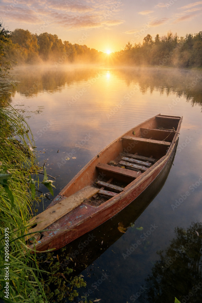 Obraz premium A wooden boat floats on a misty river at sunrise, surrounded by lush green forest trees.